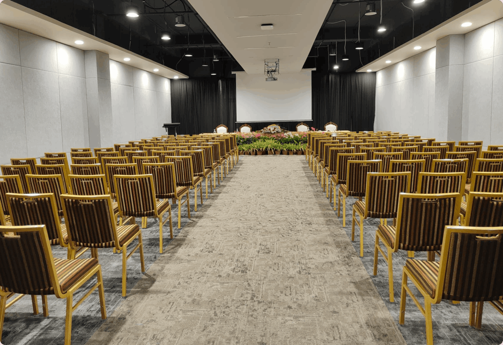 Large conference room with wooden chairs facing a stage, white screen, and floral arrangements