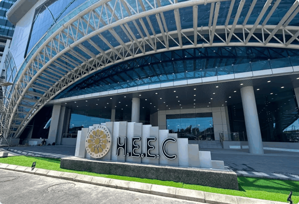 Modern glass building entrance with arched roof and "H.E.E.C." sign in landscaped foreground.