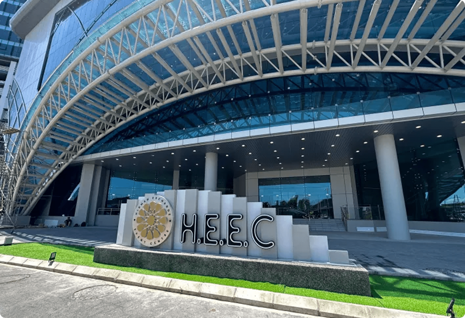 Modern glass building entrance with arched roof and "H.E.E.C." sign in landscaped foreground.